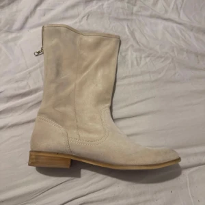 Beige mocka boots från Wojas - Snygga boots i beige mocka från Wojas med låg klack och rund tå. Skorna har träfärgad sula och dragkedja på sidan för enkel på- och avtagning. Perfekt för dig som gillar stilrena och neutrala färger. Passar dig som vill ha en klassisk look med modern touch.