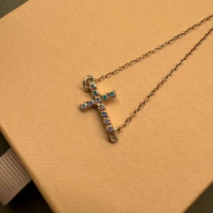 Armband med glittrigt kors i silver - Snyggt armband med ett kors dekorerat med glittrande stenar i blått, rosa och silver. Kedjan är tunn och korsets stenar ger en riktigt cool och färgglad vibe. Perfekt för dig som gillar detaljer som sticker ut och vill ha något unikt på handleden.