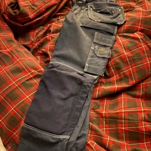 Mörkblå cargopants från Fristads Kansas - Säljer ett par mörkblå snickar/jobb byxor från Fristads Kansas med kontrastsömmar och flera praktiska fickor på benen. Byxorna är klassiska tillverkade i slitstarkt bomullsmaterial. Passar dig som är 184-192 cm lång. Ny pris 1300kr, säljer för 500kr, kom med bud så sänker vi vid snabb affär.