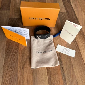Armband - Snyggt svart armband från Louis Vuitton i äkta skinn med klassiskt monogrammönster och silverfärgat LV-spänne. Armbandet har en stilren design och levereras med originalpåse, box och äkthetskort. Perfekt accessoar för att lyfta din outfit.