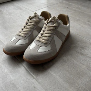 Mason margiela skor - Snygga sneakers i beige och vitt med klassisk design. Ovandelen är i en mix av mocka och skinn, med beige mockadetaljer och vita paneler. Skorna har snörning och en platt, brun gummisula med mönstrad undersida. Perfekta för dig som gillar stilrena och neutrala färger.