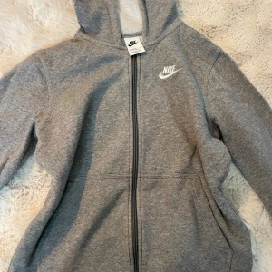 Grå Nike hoodie med dragkedja - Säljer en grå hoodie från Nike med vit logga på bröstet. Tröjan har huva, hel dragkedja framtill och två fickor. Perfekt för chill dagar eller när du vill ha en sportig look. Mjuk insida och klassisk passform.