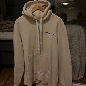 Beige hoodie från Champion - Säljer en beige hoodie från Champion med broderad logga på bröstet. Tröjan har huva med snörning, stor magficka och långa ärmar. Perfekt för chill dagar och har en clean look som funkar till det mesta. Materialet är mjuk bomull för extra komfort.