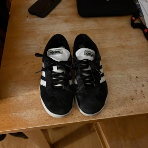 Svarta Adidas sneakers med vita ränder - Säljer ett par klassiska svarta Adidas sneakers med vita ränder på sidorna och vit sula. Skorna har snörning och är tillverkade i mocka med detaljer i syntet. Perfekta för dig som gillar en tidlös och sportig stil 💕