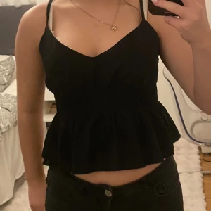 Svart croppad linnetopp med volang - Säljer en svart croppad linnetopp med smala axelband och v-ringad hals. Toppen har en smockad midja som ger en snygg passform och en volang nedtill. 