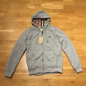 Burberry zip hoodie - Grå zip hoodie från Burberry med logga på bröstet och klassisk huva med snörning. Tröjan har dragkedja framtill, och i är mjukt bomullsmaterial. Hoodien är i storlek S och är i nyskick hör av er vid frågor
