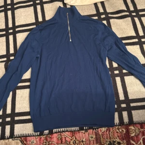 Blå merinoull polotröja från Riley - Snygg blå polotröja från Riley i mjuk merinoull. Tröjan har en hög krage med half zip-dragkedja och långa ärmar. Perfekt för lager-på-lager och chill stil under höst och vinter.