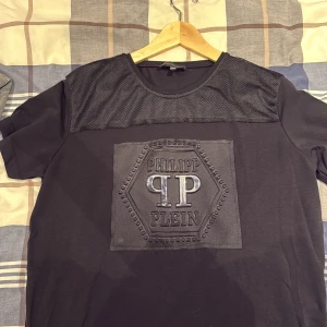 Svart Philipp Plein t-shirt med mesh och dragkedjor - Svart t-shirt från Philipp Plein i storlek XL med coolt meshparti över axlarna och ett stort, upphöjt PP-logomärke framtill. Snygga guldfärgade dragkedjor nertill ger extra edge. Passar dig som gillar street och exklusiv stil.