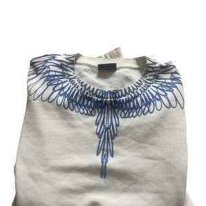 Vit sweatshirt från Marcelo Burlon - Vit sweatshirt från Marcelo Burlon med blått grafiskt mönster runt halsen som liknar fjädrar. Tröjan har rund hals och är tillverkad i mjuk bomull. Perfekt för dig som gillar streetwear och vill sticka ut med cool design. OBS har ett litet hål på baksidan men det är så litet att det inte märks. Pris kan diskuteras vid smidigaffär. Sitter perfekt på mig som är 180 cm!