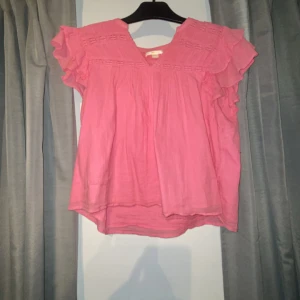 Rosa blus med volangärmar - Säljer en söt rosa blus med volangdetaljer på ärmarna och v-ringad hals. Blusen har ett luftigt och flowy material, perfekt för dig som gillar en feminin stil. Dekorativa sömmar upptill ger extra charm. Passar dig som vill ha en chill och snygg look.