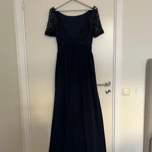 Marinblå långklänning med spets - Elegant marinblå långklänning med v-ringning i ryggen och korta ärmar i blommig spets. Kjolen är mjukt fallande och markerad midja ger en snygg siluett. Perfekt för bal eller festliga tillfällen. Den är aldrig använd.