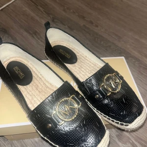 Svarta espadrillos Michael Kors - Snygga svarta espadrillos från Michael Kors med präglat mönster och guldfärgad MK-logga framtill. Skorna har flätad sula i naturmaterial och detaljer i svart skinnliknande material. Perfekta för en trendig och avslappnad look.