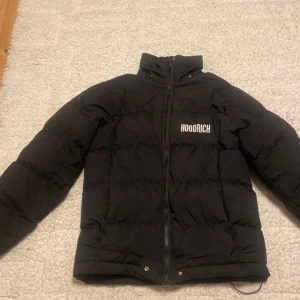 Svart pufferjacka från Hoodrich S - Svart pufferjacka från Hoodrich med broderad logga på bröstet. Jackan har hög krage, hel dragkedja framtill och quiltad design. Materialet är syntet och polyester, perfekt för kalla dagar. Snygg och stilren look med loose passform.