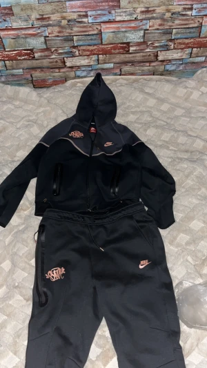 Central Cee Tracksuit Nike Tech - Nike tracksuit i svart med ljusrosa detaljer och loggor. Hoodien har dragkedja, två fickor och snyggt tryck på bröstet. Byxorna har resår i midjan och matchande logga. Skön passform och sportig look, perfekt för chill eller träning.