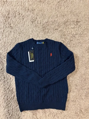 Marinblå stickad tröja Polo Ralph Lauren - Snygg marinblå stickad tröja från Polo Ralph Lauren med klassisk kabelstickning och röd broderad logga på bröstet. Rund hals och långa ärmar, perfekt för dig som gillar stilrena och tidlösa plagg. Skriv vid någon fundering ✅