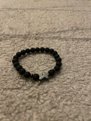 Svart pärlarmband med kors - Stilrent svart pärlarmband med runda matta pärlor och ett metalliskt kors i silverfärg som detalj. Armbandet har elastisk tråd och passar de flesta handleder. Perfekt för dig som gillar minimalistisk och cool design.