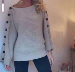 Beige stickad tröja med knappar - Snygg beige stickad tröja med bred halsringning och svarta knappar längs hela ärmarna. Tröjan har lång ärm och en loose passform som ger en chill vibe. Perfekt att matcha med jeans eller leggings för en enkel och stilren look.