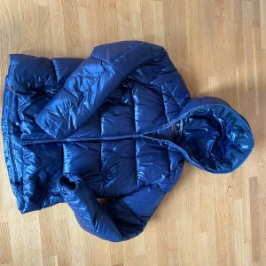 Blå pufferjacka från Save The Duck - Säljer en blå pufferjacka från Save The Duck i storlek S. Jackan är 100% animal free, har huva, dragkedja framtill och två fickor med dragkedja. Materialet är glansigt och syntetiskt, perfekt för kalla dagar. Snygg och modern look med quiltad design. Grisch som fan och skickar samma dag du beställer 