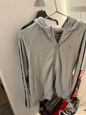 Grå hoodie Tommy Hilfiger med dragkedja - Snygg grå hoodie från Tommy Hilfiger med huva och hel dragkedja. Långärmad med marinblå, vit och röd rand längs ärmarna, samt stort Hilfiger-tryck på ena ärmen. Mjuk bomullskänsla och klassisk logga på bröstet. Perfekt för chill eller streetwear.
