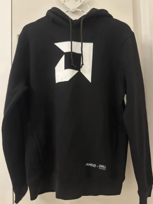 Svart AMD x Dell hoodie med tryck - Svart hoodie med stor vit AMD-logga på bröstet och AMD x Dell Technologies-tryck fram och bak. Tröjan har huva med snörning och är tillverkad av 80% bomull och 20% återvunnen polyester. Perfekt för dig som gillar tech och vill ha en clean look.