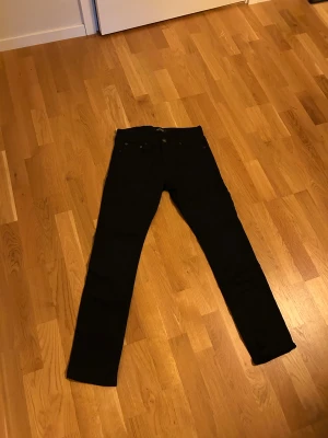 Svarta Slim Glenn jeans från Jack & Jones - Svarta jeans från Jack & Jones, modell Slim Glenn. Snygg slim fit med klassisk femficksdesign och knappstängning. Jeansen är tillverkade i ett stretchigt bomullsmaterial som sitter tajt men bekvämt längs benen. Perfekt för dig som gillar en stilren och modern look.