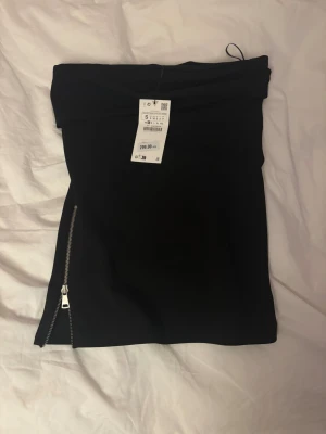 Svart tubtopp med dragkedja från Zara  - Svart tight tubtoppl från Zara med silvriga dragkedjor på sidorna. Toppen har en enkel och stilren design som passar perfekt till en trendig look. Gjord i stretchigt material för en snygg passform.