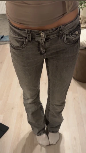 Grå bootcut jeans med låg midja - Säljer ett par grå jeans med låg midja och bootcut-ben. Jeansen har klassiska fickor fram och bak, samt extra fickdetalj med knapp bak. Perfekta för dig som gillar en avslappnad men trendig look.