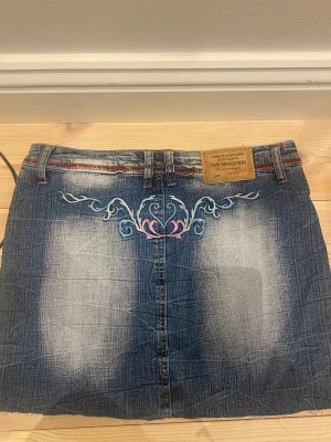 Broderad jeanskjol från New Sensation - Snygg kort jeanskjol från New Sensation med slitna detaljer och broderier i blått, vitt och rosa. Kjolen har bälteshällor, röda kontrastsömmar och broderat hjärta samt dekorativa mönster både fram och bak. Perfekt för dig som gillar unik denimstil.