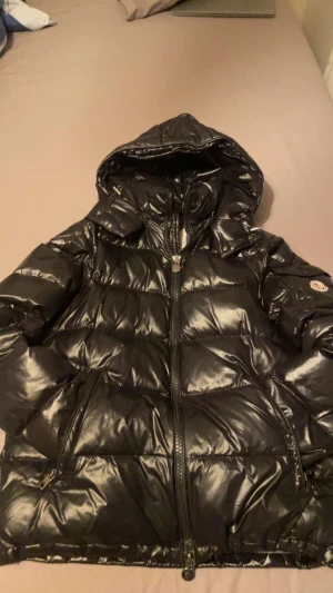 Svart Moncler pufferjacka med huva - Svart pufferjacka från Moncler med glansig finish, huva och två sidofickor med dragkedja. Jackan har quiltad design, lång ärm och klassisk Moncler-logga på ärmen. Insidan har ett unikt serietidningsmärke. Perfekt för kalla dagar.