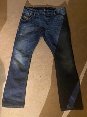 Diesel blå jeans straight fit - Säljer ett par Diesel jeans i klassisk blå tvätt med snygga slitningar och kontrastsömmar. Modellen har raka ben, fem fickor och detaljerade bakfickor med gul söm. Midjemått:43 cm och innerbenslängd 73 cm.