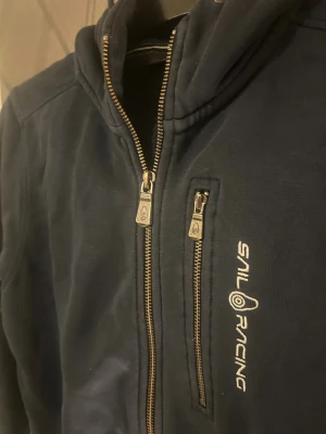 Marinblå hoodie från Sail Racing - Marinblå hoodie från Sail Racing med dragkedja framtill och på bröstfickan. Tröjan har huva och broderad logga på bröstet. Perfekt för dig som gillar sportig och avslappnad stil.
