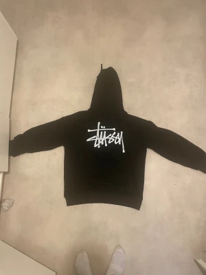 Svart Stüssy hoodie med logga - Svart hoodie från Stüssy med stor vit logga på ryggen och mindre logga på bröstet. Klassisk modell med huva och långa ärmar, perfekt för en streetwear-look. Mjuk och skön, 