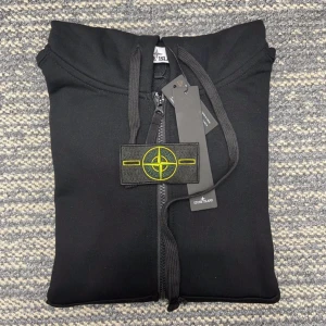 Svart hoodie från Stone Island - Svart hoodie från Stone Island med dragkedja framtill och klassisk logopatch på vänster ärm. Tröjan har huva med snörning och två fickor fram. Materialet är mjuk bomull och passformen är normal. Perfekt för dig som gillar streetwear och snygga detaljer.