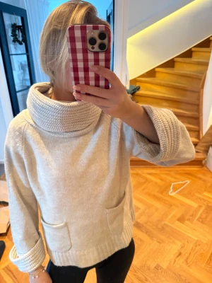 Beige stickad polotröja med fickor - Mysig beige stickad polotröja med bred, ribbad krage och uppvikta ärmslut. Tröjan har två praktiska fickor framtill och en relaxed passform som är perfekt för chill dagar. Passar dig som gillar enkel och stilren design.