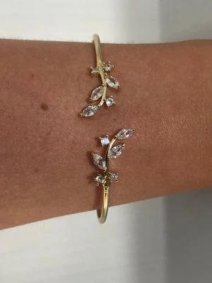 Guldigt armband med kristaller - Snyggt guldigt armband med glittrande kristaller i form av blad och små stenar. Designen är elegant och har en tunn, böjd form som sitter fint runt handleden. Perfekt för dig som gillar detaljer och vill ha något som sticker ut.