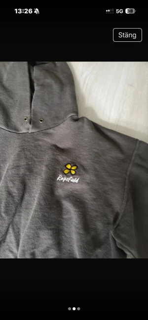 Mörkgrå hoodie från Rapsfield - Säljer en mörkgrå hoodie från Rapsfield med broderad gul blomma och vit logga på bröstet. Tröjan har huva med snörningshål, magficka och ribbade muddar vid ärmslut och nederkant. Perfekt för chill dagar och sköna outfits.