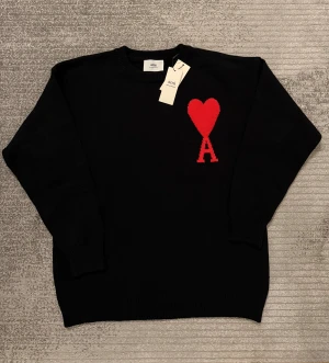 Svart stickad tröja från Ami Paris - Svart stickad tröja från Ami Paris med rött hjärta och bokstaven A broderat på bröstet. Tröjan har rund hals, ribbade muddar och lång ärm. Perfekt för dig som gillar stilrena detaljer och vill ha något som sticker ut.