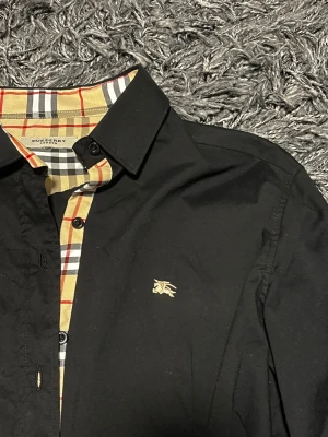 Svart Burberry skjorta med rutiga detaljer - Svart skjorta från Burberry med klassiska rutiga detaljer i beige, rött, vitt och svart på insidan av kragen, knappslån och ärmsluten. Skjortan har lång ärm, knappar framtill och ikonisk broderad logga på bröstet. Snygg och tidlös design i bomull.