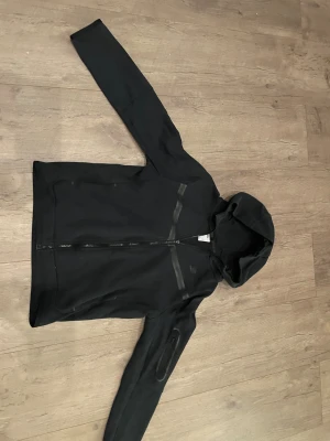Svart Nike Tech tröja i storlek 170 - Svart Nike tech från nike i storlek 158-170. Skön tröja som jag använt mycket men den är fortfarande i bra skick. Har också råkat göra sönder själva zip upen. Men den går fortfarande bra att dra upp. Mitt pris är 250 kr!