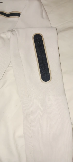 Vit hoodie från Nike med dragkedja XL - Nike hoodie i vit med svarta och beige detaljer, hel dragkedja framtill och huva. Svart Nike-logga på bröstet och svarta ränder över bröstet och ärmslut. Praktisk ficka med dragkedja på ärmen. Standard fit, mjukt bomullsmaterial.