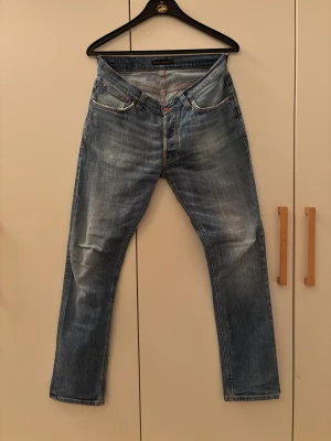 Nudie Jeans ljusblå jeans 31/32 - Säljer ett par ljusblå jeans från Nudie Jeans i klassisk femficksmodell. Jeansen har raka ben, orangea sömmar och snygga slitningar framtill och baktill. Materialet är 100% bomull och passformen är normal. Perfekta för dig som gillar en avslappnad stil.