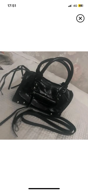 Svart handväska med dragkedja och nitar - Cool svart handväska i glansigt skinnliknande material med silverfärgade detaljer, dragkedja framtill och dekorativa nitar. Väskan har både handtag och avtagbar axelrem samt långa snören på sidorna för extra edge. Perfekt storlek för det viktigaste.