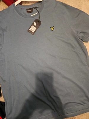 Blå t-shirt från Lyle & Scott - Snygg blå t-shirt från Lyle & Scott med klassisk rund hals och korta ärmar. På bröstet sitter den ikoniska gula fågeln som broderad detalj. T-shirten är gjord i mjuk bomull och har en clean, stilren look som funkar till allt.