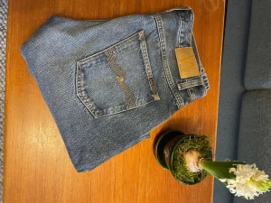 Blå jeans från Nudie Jeans Co, 32/32 - Snygga blå jeans från Nudie Jeans Co i klassisk straight fit. Jeansen har en cool tvätt med slitningar och Nudie’s ikoniska broderade bakfickor. Materialet är robust denim i bomull och passformen är normal med raka ben. Perfekt för dig som gillar tidlös stil och streetwear.