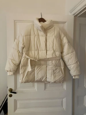 Cremevit/vit pufferjacka/täckjacka med bälte - Säljer en vit pufferjacka med hög krage och quiltad design. Jackan har ett matchande bälte i midjan och långa ärmar med ribbade muddar. Perfekt för kalla dagar och har en oversized passform som ger en cool och avslappnad look. (Har en likadan själv som jag använt mycket förra vintern, och kommer även använda denna tror jag😇😍. Och då jag klantigt nog råkade klicka i min beställning två gånger så fick jag hem två🫣😅).  Helt ny oanvänd och bara upptagen ur förpackningen för foton. 🌸