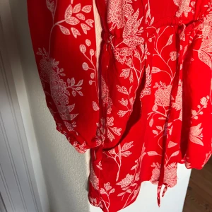Röd shortsdress H&M - Superfin röd jumpsuit /shorts från H&M med vitt blommigt mönster, v-ringning med osynlig knäppning fram och långa ärmar. Resår och tunt knytband i midjan. Cool modell med praktiska fickor. Skräddad i ett lätt och skönt mjukt material. Perfekt för sommarhalvåret både med eller utan strumpbyxor. 