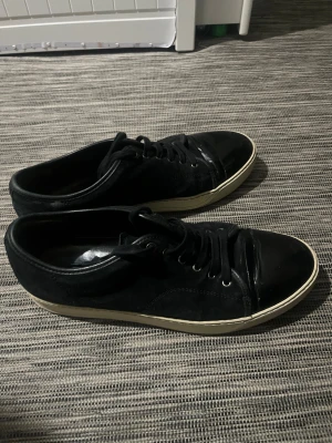 Svarta sneakers från Lanvin - Svarta sneakers från Lanvin med snygg kontrast mellan mocka och blankt skinn på tån. Klassisk rund tå, svart snörning och vit platt sula. Perfekt för dig som gillar stilrena och lyxiga detaljer i din vardag. Märket syns på sulan. Tyvärr så är boxen borta 