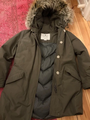 Olivgrön parkas från Woolrich med päls - Säljer en olivgrön parkas från Woolrich med snygg päls på huvan. Jackan har långa ärmar, dragkedja och knappar framtill, samt två stora fickor. Insidan är quiltad och fodrad för extra värme. Perfekt för kalla vinterdagar och har en klassisk, clean look.