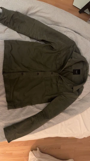 Olivgrön overshirt från New Look Men - Olivgrön overshirt från New Look Men med klassisk krage, två bröstfickor med lock och knäppning framtill. Skjortan är i bomull och har lång ärm. Perfekt lager-på-lager-plagg med clean look och raka linjer.