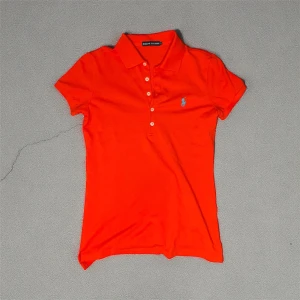 Orange pikétröja från Ralph Lauren, Stl S, i ny skick  - Snygg orange pikétröja från Ralph Lauren i storlek S. B 35cm L 61cm. Klassisk krage, korta ärmar och knäppning med vita knappar framtill. Tröjan har det ikoniska blå broderade Polo-logan på bröstet och är gjord i mjuk bommul. I nytt skick. Kommer från djur-rökfritt hem